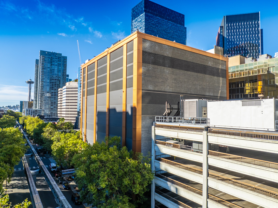 Urban landscape of Seattle featuring a large orange and gray building in the foreground, with skyscrapers and the Space Needle visible in the background.