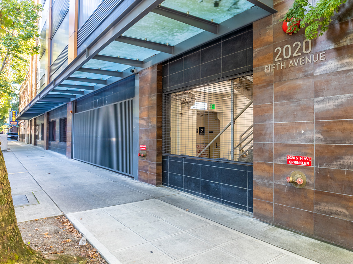 Street-level view of a modern building entrance at 2020 Fifth Avenue in Seattle, featuring glass canopy, metal grating, and brown tiled exterior.