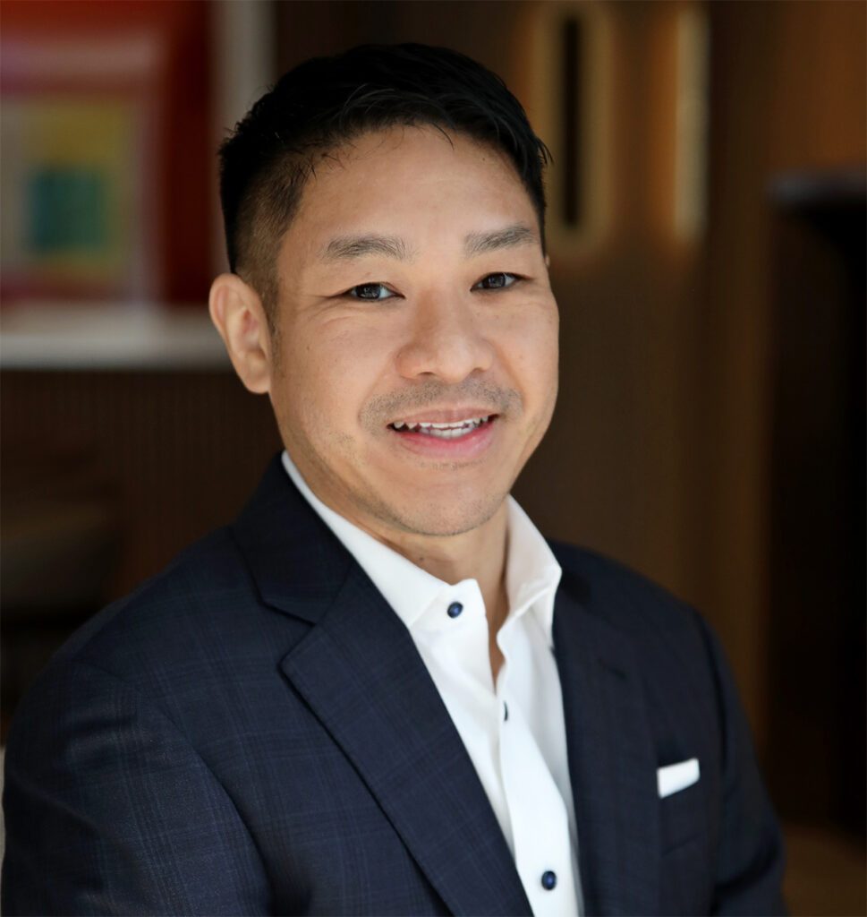 Smiling man with short dark hair wearing a navy suit jacket and white shirt, photographed indoors with a blurred background.