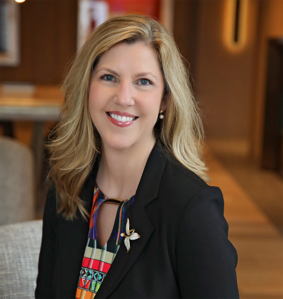 Smiling woman with long blonde hair wearing a dark blazer, photographed indoors with blurred background.