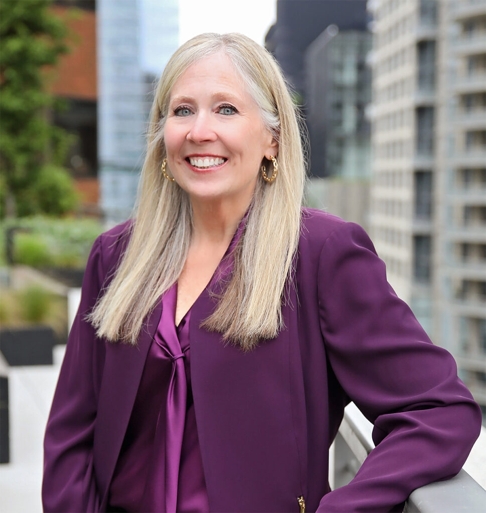 Smiling woman with long blonde hair wearing a purple blazer, photographed outdoors with city buildings in the background.