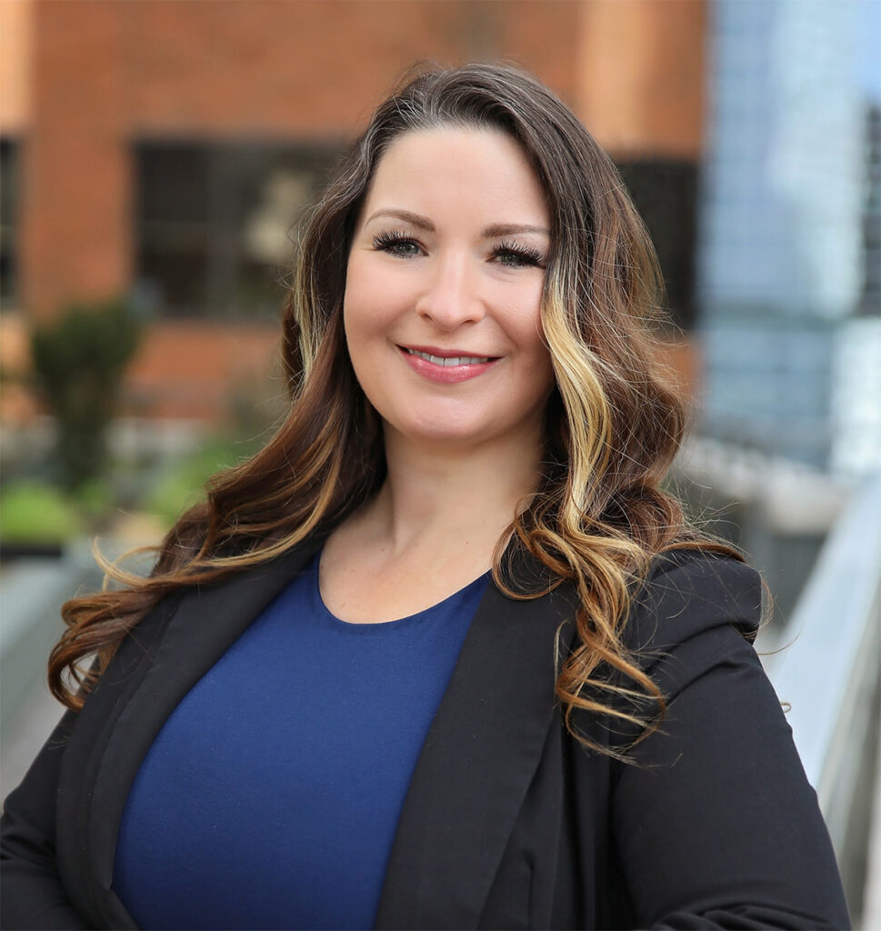 Smiling woman with long brown hair wearing a dark blazer and blue shirt, photographed outdoors with city buildings in the background.