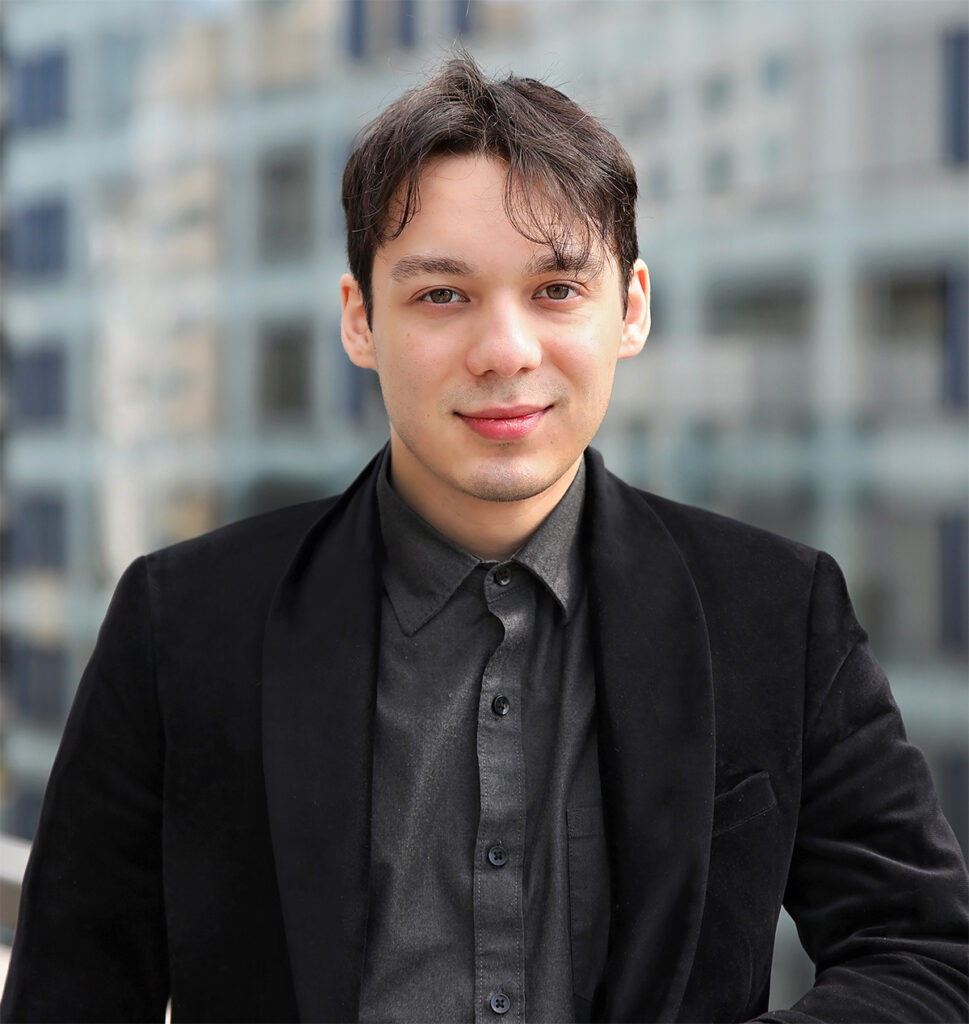 Young man with short dark hair and a slight smile, wearing black blazer and collared shirt, with blurred city buildings in the background.