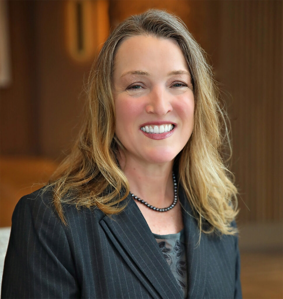 Smiling woman with shoulder-length brown hair wearing dark blazer and blouse, indoors.