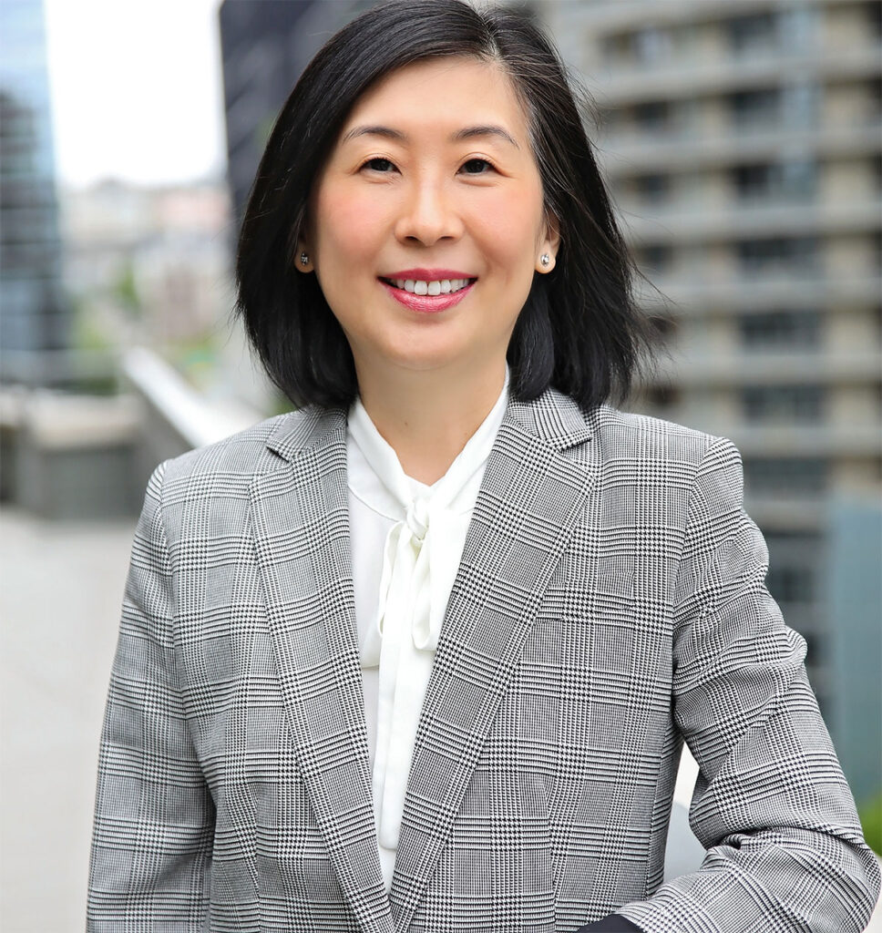 Smiling Asian woman with shoulder-length black hair wearing a grey blazer and white blouse outside with blurred buildings in the background.